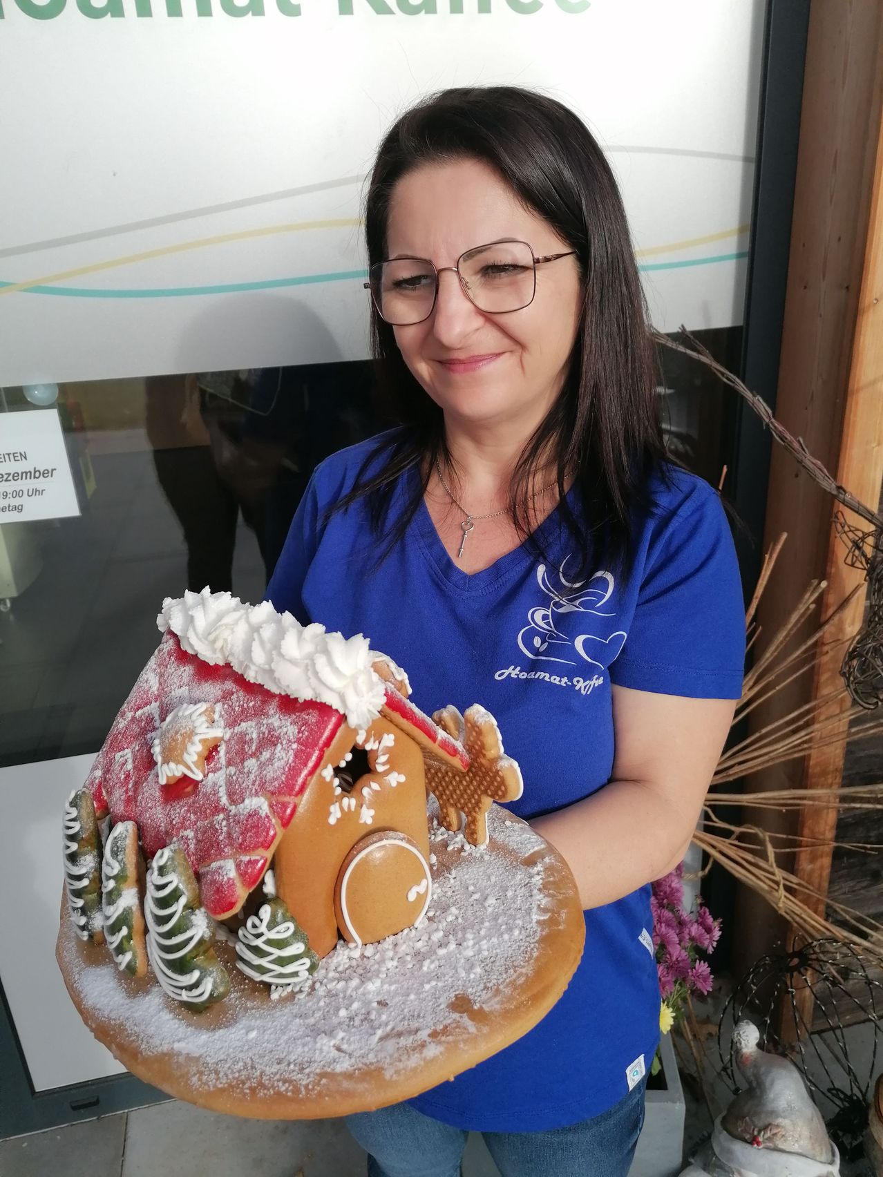 A woman wearing a blue shirt holds a gingerbread house with snow on top, standing in front of a shop.