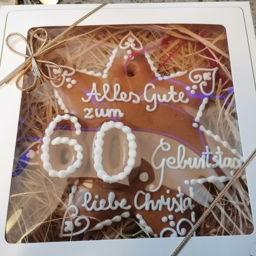 A gingerbread star in a white box, decorated with 'Alles Gute zum 60. Geburtstag liebe Christa' in white icing. It rests on hay with a gold ribbon and a silver chain.