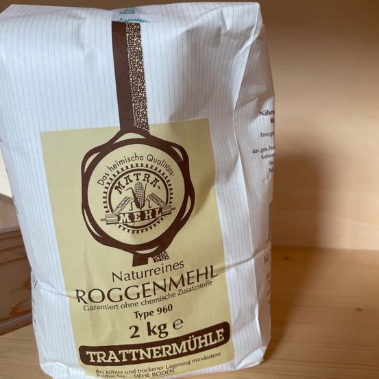 Ein weißer Sack 'Trattnermuehle Naturreines Roggenmehl', eine Marke für Roggenmehl, mit einem Gewicht von 2 kg, und einem gelben Etikett mit Logo und Text auf Deutsch. Das Etikett zeigt an, dass es garantiert ohne chemische Zusätze ist.