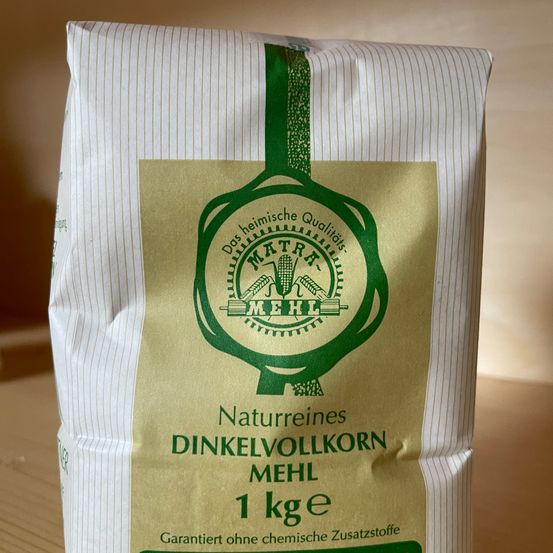 Ein Beutel Mehl-Marken-Natur-Vollkorn-Dinkel mit einem Gewicht von 1 kg. Es ist frei von chemischen Zusatzstoffen. Die Verpackung ist beige mit grünen Akzenten.