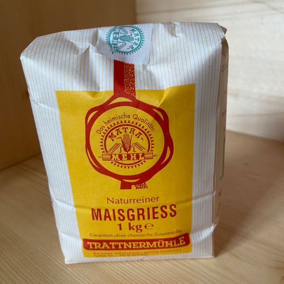 Ein Beutel 'Naturreiner Maisgrieß' wird gezeigt. Es ist ein gelber und weißer Beutel mit einem Gewicht von 1 kg. Er ist garantiert ohne chemische Zusatzstoffe.