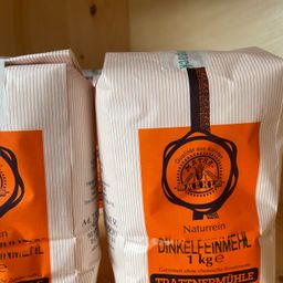 Zwei Säcke Dinkelfeinmeh, ein Naturprodukt mit 1kg, auf einem hölzernen Hintergrund ausgestellt. Die Säcke haben orangefarbene Etiketten mit dem Markennamen und den Produktdetails.
