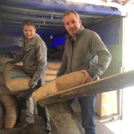 Zwei Männer entladen einen Lkw voller Zementsäcke. Ein Mann hält einen Sack, während der andere einen großen Sack auf einer Holzplatte balanciert.