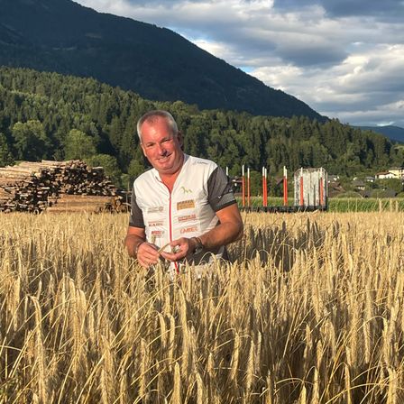 Ein Mann in einem weißen Shirt steht in einem goldenen Weizenfeld und lächelt in die Kamera. Hinter ihm befinden sich Berge, Bäume und ein Holzstapel.