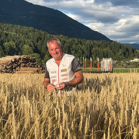 Ein Mann in einem weißen Shirt steht in einem goldenen Weizenfeld und lächelt in die Kamera. Hinter ihm befinden sich Berge, Bäume und ein Holzstapel.