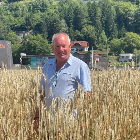 Ein Mann steht in einem Weizenfeld mit einem Wald und Häusern im Hintergrund.