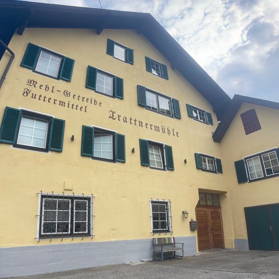 Ein gelbes Gebäude mit grünen Fensterläden trägt ein Schild mit der Aufschrift 'Mehri-Betreide Futtermittel Trattnermuhle'. Es gibt mehrere Fenster, eine Bank und eine Lampe draußen. Der Himmel ist bewölkt.
