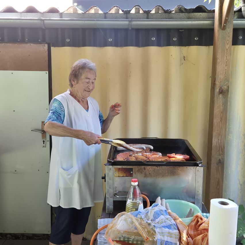 Eine ältere Frau grillt Essen auf einem Metallgrill im Garten. Sie trägt ein weißes Schürzen und hat ein lächelndes Gesicht. Es gibt eine Flasche Öl und eine Schüssel auf dem Tisch.