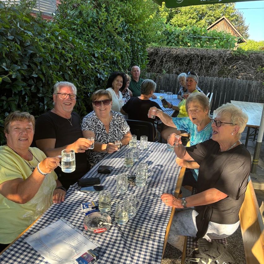 Eine Gruppe von Erwachsenen sitzt an einem Tisch im Freien, einige lächeln und halten Gläser. Sie sind von Grün umgeben, und es gibt Stühle und einen Tisch hinter ihnen.