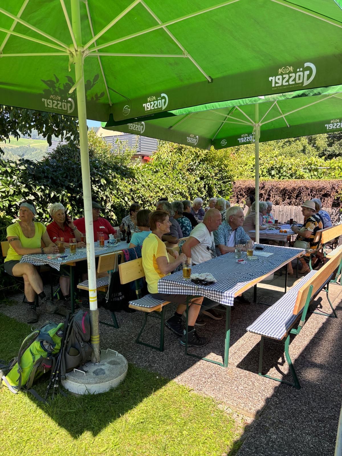 Eine Gruppe von Menschen sitzt unter grünen Sonnenschirmen an Tischen in einem Außenrestaurant, umgeben von Grünpflanzen und Bäumen. Sie führen Gespräche, und einige halten Getränke in den Händen.