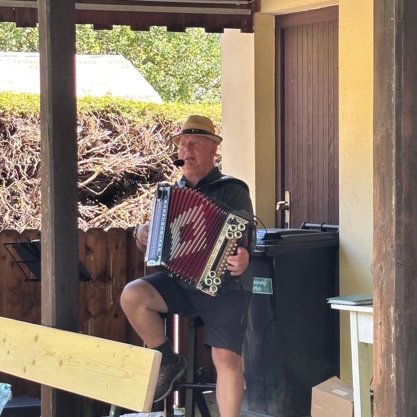 Ein Mann mit Hut spielt Akkordeon sitzend auf einem Stuhl im Freien. Hinter ihm ist ein Holzzaun und eine Tür zu sehen.