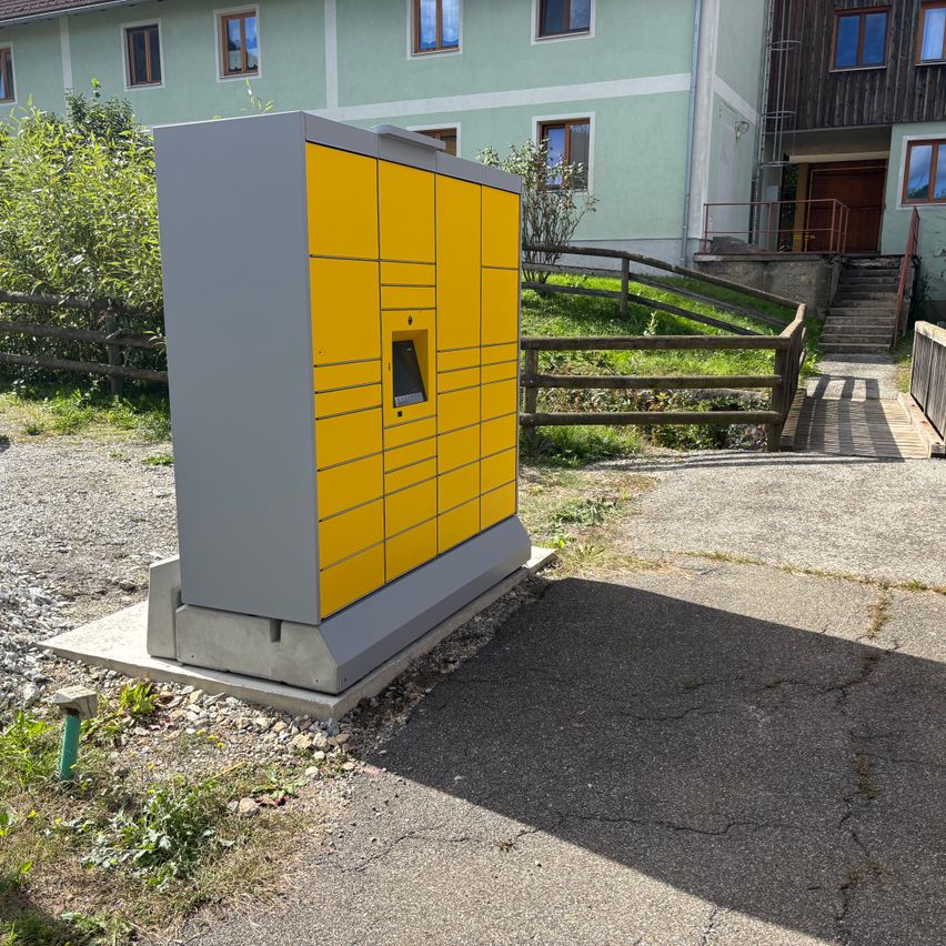 Ein gelber und grauer Paketschließfachkasten ist vor einem grünen Gebäude mit Treppen auf einem Betonsockel installiert.