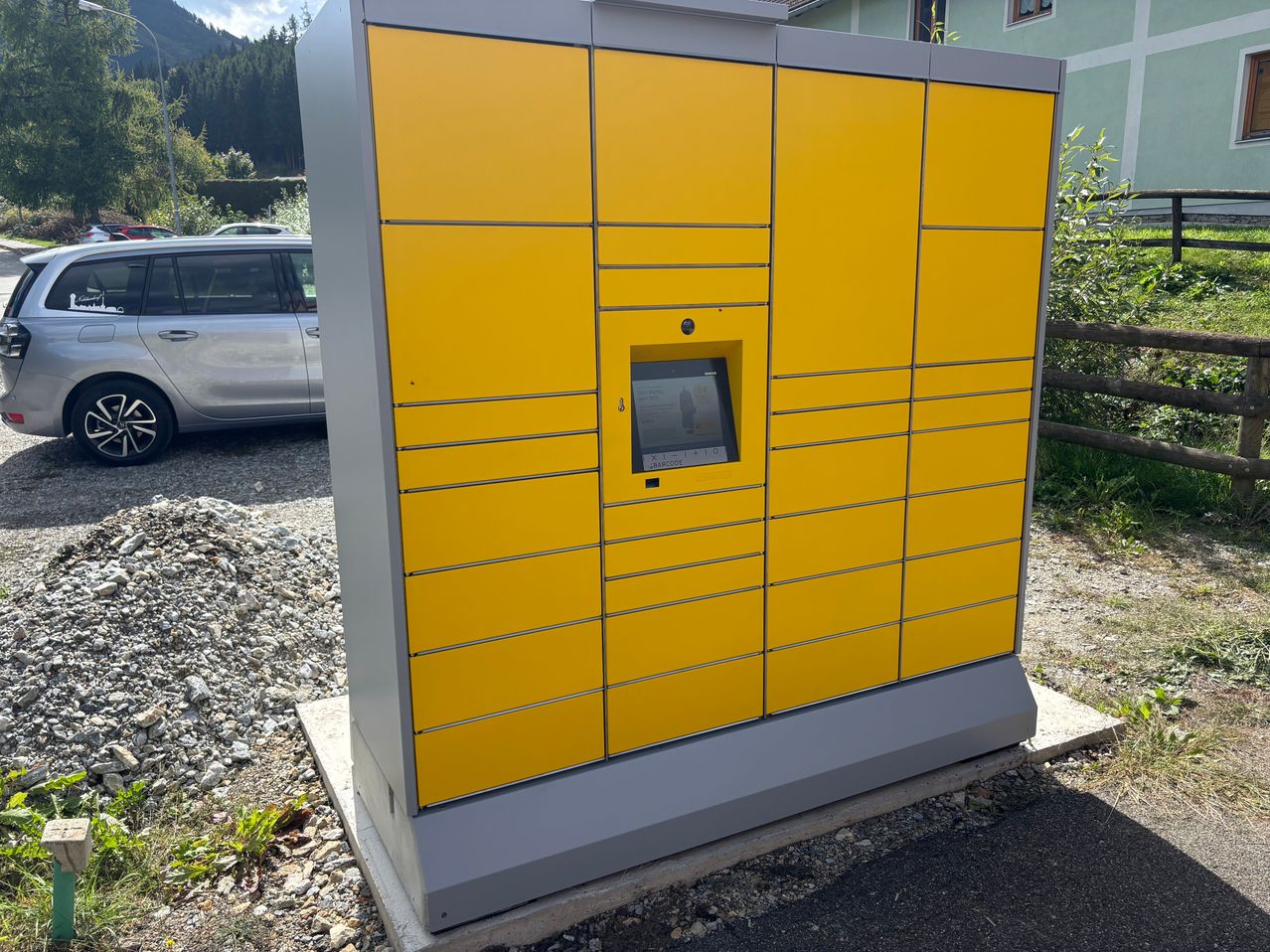 Ein gelber Paketschrank ist an der Straßenseite installiert. Er hat einen Bildschirm und eine Tastatur an der Tür. Ein Auto ist in der Nähe geparkt.