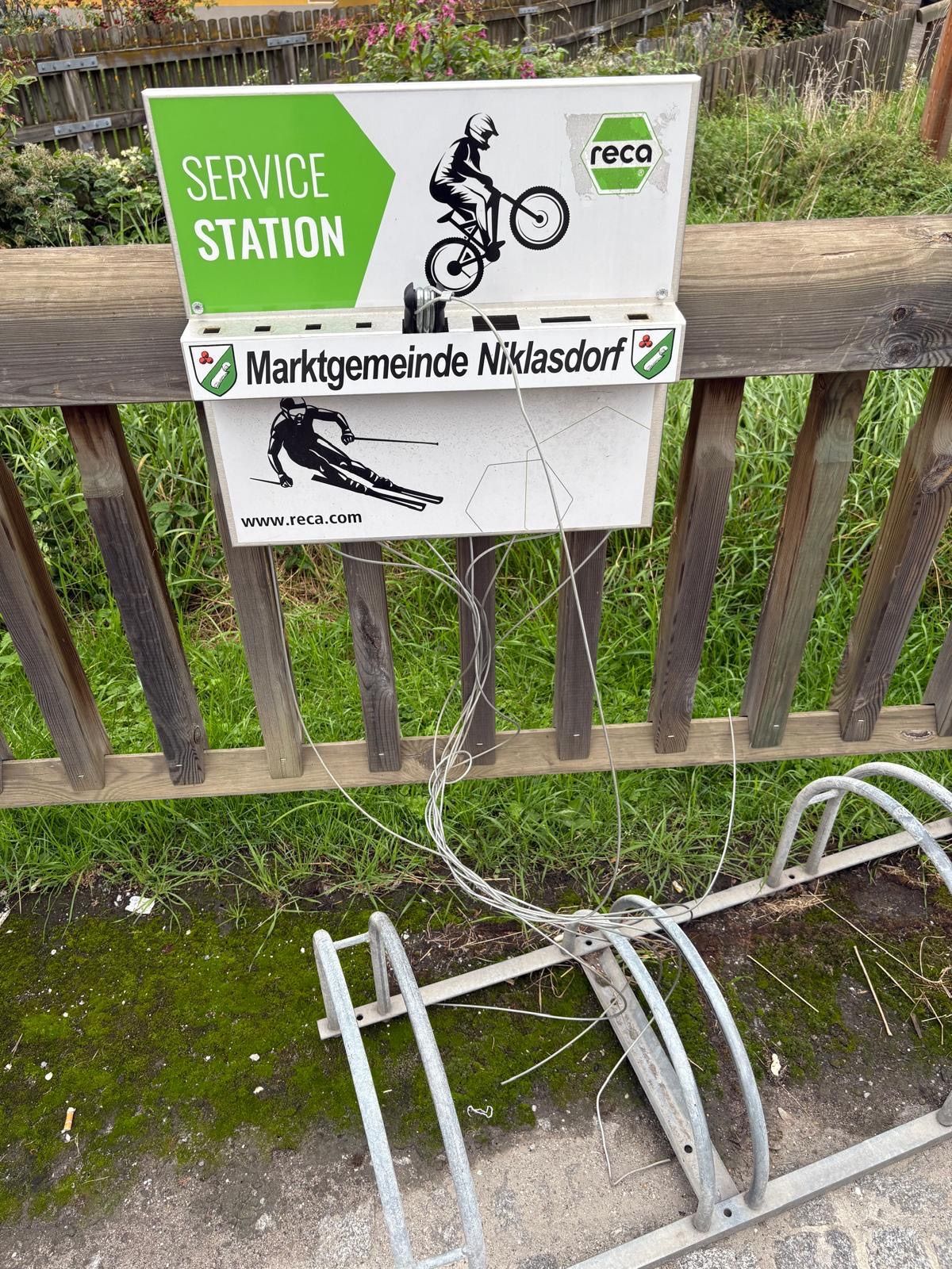 Ein Schild für eine Servicestation hängt an einem Holzzaun, mit einem Fahrradbild darauf. Darunter befindet sich ein Schild für Marktgemeinde Niklasdorf mit einem Skifahrer und einer Website-Adresse.