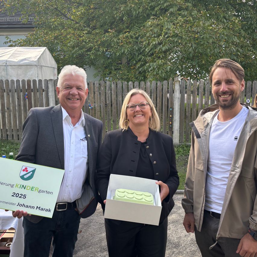 Drei Menschen stehen lächelnd draußen. Ein Mann hält ein Schild für Kindergarten 2025. Eine Frau hält eine Box mit einem grünen Lego-Kuchen. Sie stehen in der Nähe eines Holzzauns und Bäumen.