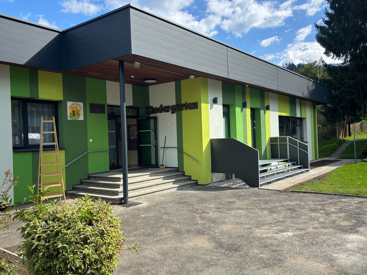 Der Eingang eines Kindergartens mit grün-weiß gestreiften Wänden. Es gibt Treppen, die zum Eingang führen, mit einem Handlauf an der Seite. Eine Leiter ist auf der linken Seite platziert.
