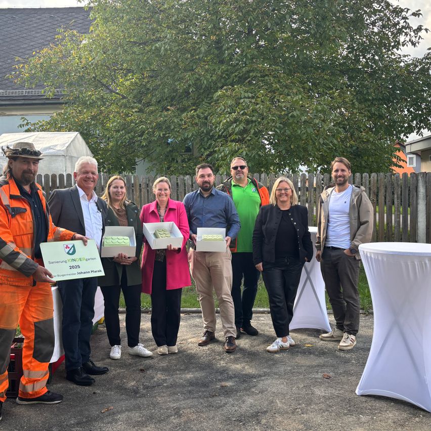 Eine Gruppe von Menschen steht vor einem Haus, einige halten Geschenkboxen. Die Person auf der linken Seite trägt einen Hut und hält ein Banner. Bäume und ein Zaun sind im Hintergrund.