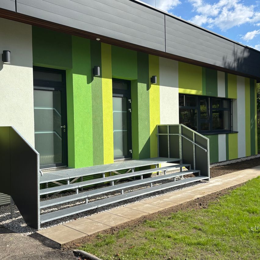 Ein modernes Gebäude mit grün-gelb gestreifter Außenfassade. Es verfügt über eine Metalltreppe mit Handläufen, zwei Türen und zwei an der Wand montierten Lampen. Das Gebäude hat ein Fenster mit schwarzem Rahmen und einem weißen Dach.