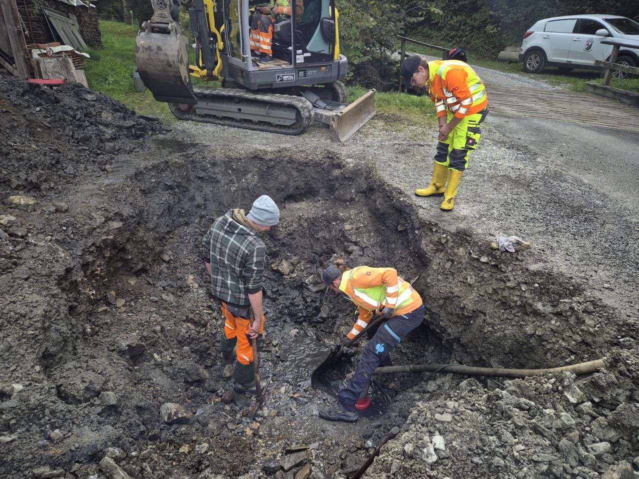 Drei Arbeiter graben in einer schlammigen Grube, einer in einem Loch, ein anderer daneben. Ein gelber Bagger ist in der Nähe geparkt. Ein Auto ist auf der Straße im Hintergrund geparkt.
