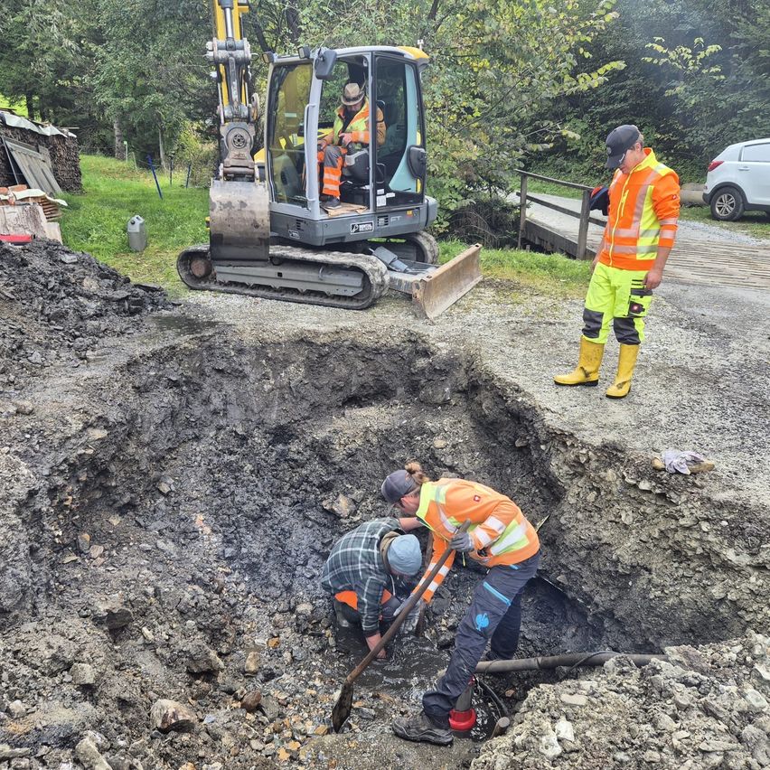Bauarbeiter graben ein Loch, während ein Mann in reflektierender Kleidung zusieht. Ein Baufahrzeug ist in der Nähe.
