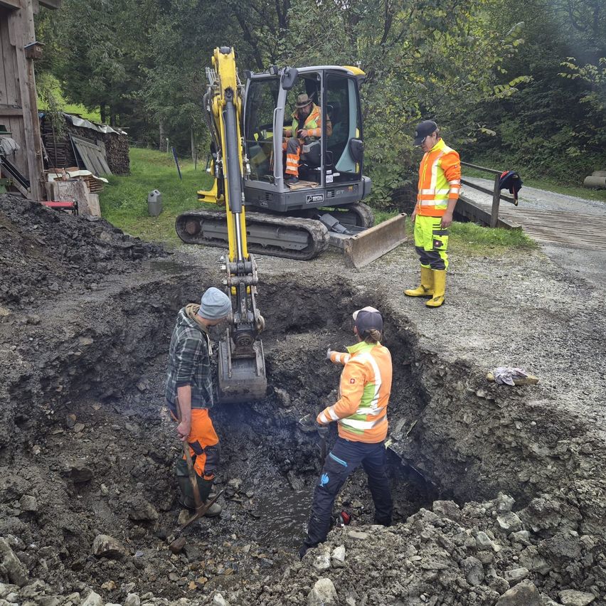 Mehrere Arbeiter sind auf einer Baustelle mit einem großen Loch, einer bedient einen gelben Bagger. Hinter ihnen beobachtet ein Mann in reflektierender Kleidung.
