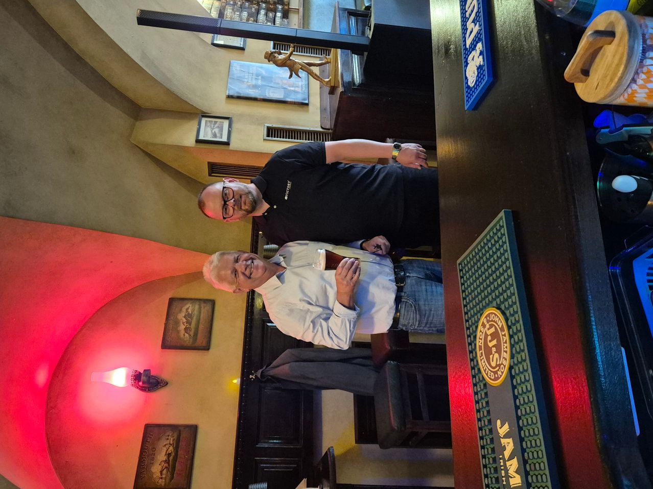 Zwei Männer posieren für ein Foto in einer Bar. Ein Mann hält ein Glas, während der andere eine Flasche hält. Es gibt ein Schild, das JJ's steht. Es gibt auch einige Bilderrahmen, die an der Wand hängen.