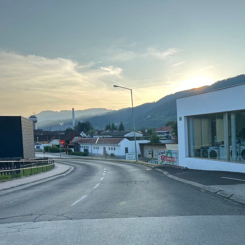 Eine leere Straße bei Sonnenuntergang mit Bergen und Häusern im Hintergrund. Rechts ist ein Gebäude mit sichtbaren Waschmaschinen zu sehen.