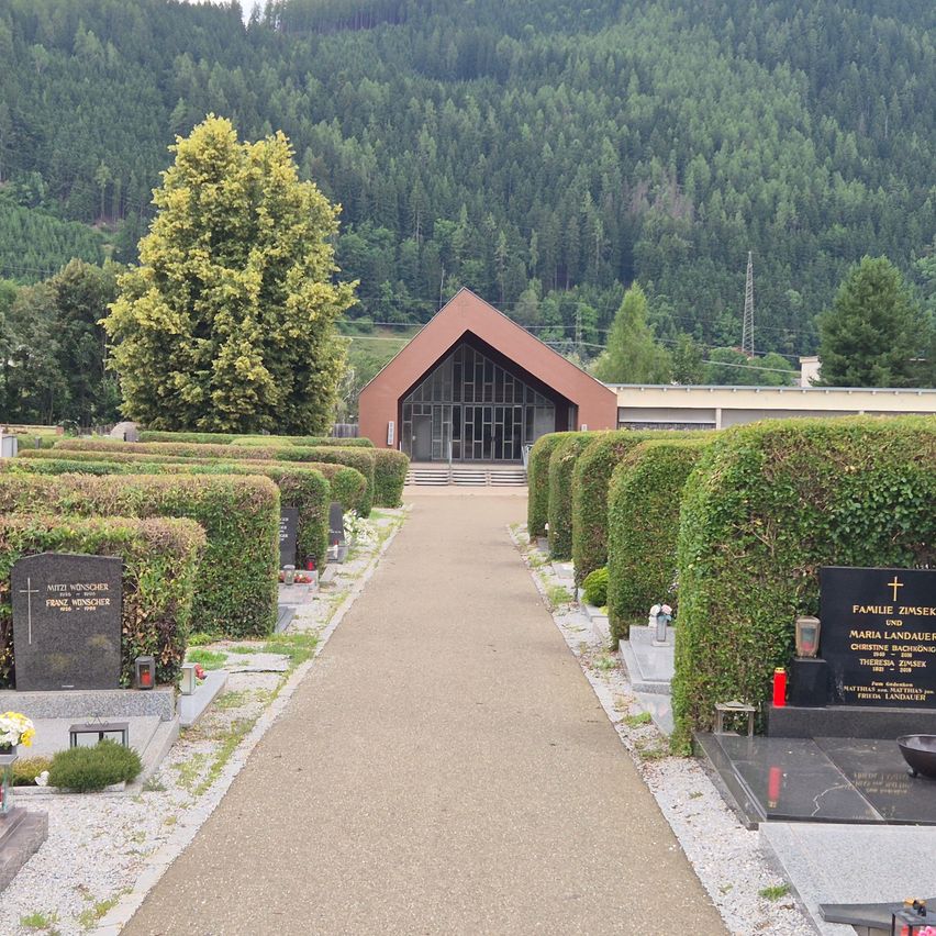Ein Friedhof mit Grabsteinen, Hecken und einer Kapelle mit Glasfenstern. Ein Weg führt zur Kapelle, und es gibt eine Aussicht auf einen Berg in der Ferne.