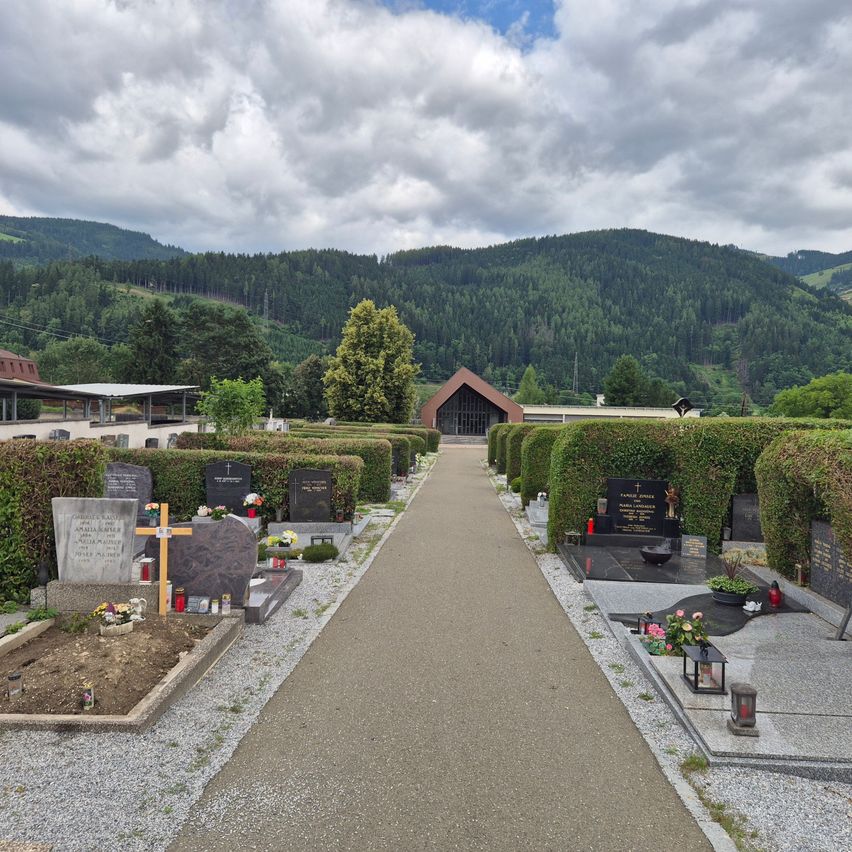 Ein ruhiger Friedhof mit ordentlich angeordneten Grabsteinen, umgeben von Hecken, mit einem Weg, der zu einer Kapelle führt, vor dem Hintergrund von Bergen.