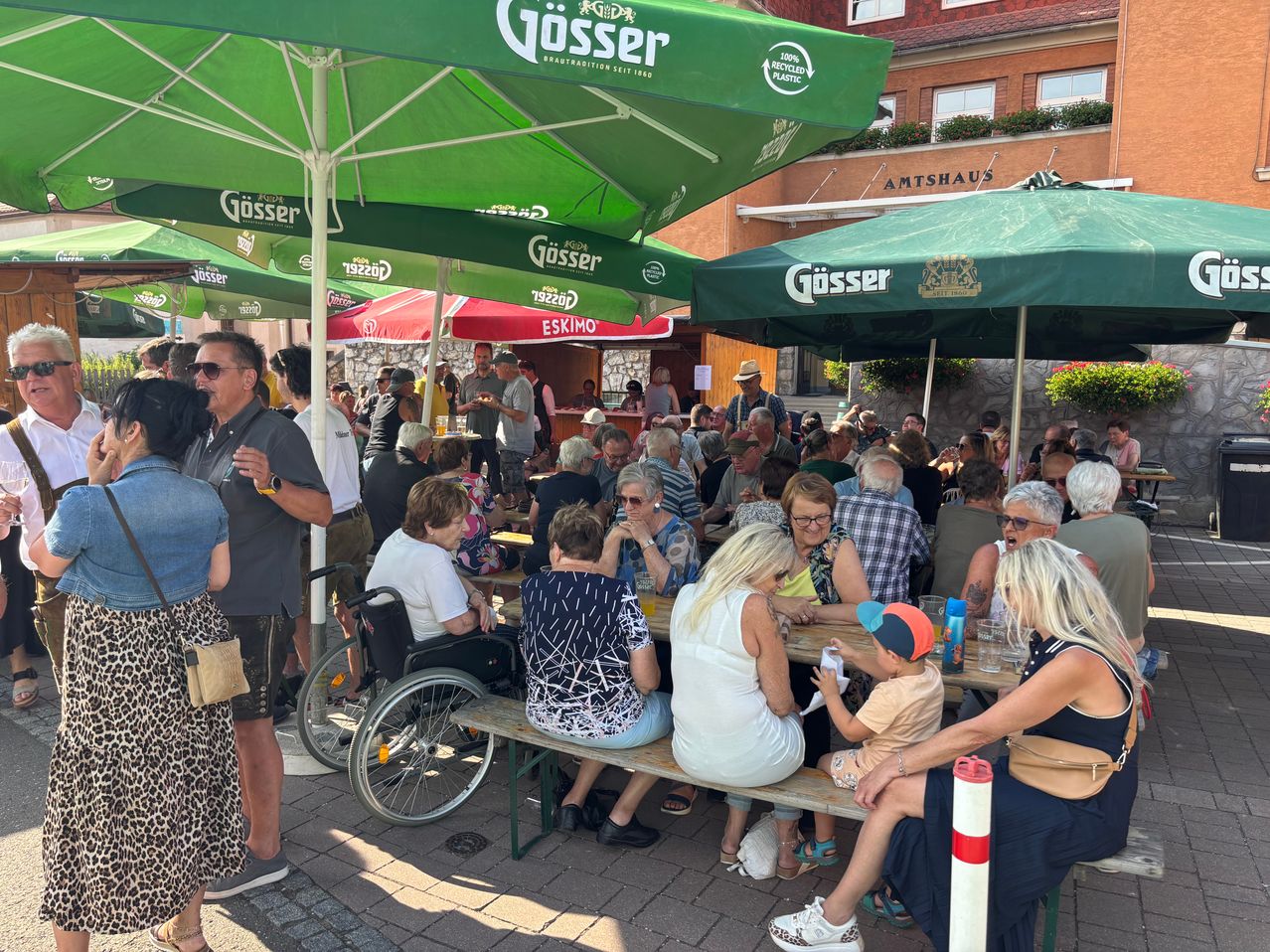 Eine Menschenmenge versammelt sich in einem Außenrestaurant mit grünen Schirmen, einige sitzen, während andere stehen und plaudern. Ein Mann im Rollstuhl ist anwesend, und die Tische sind mit Getränken und Essen gedeckt.