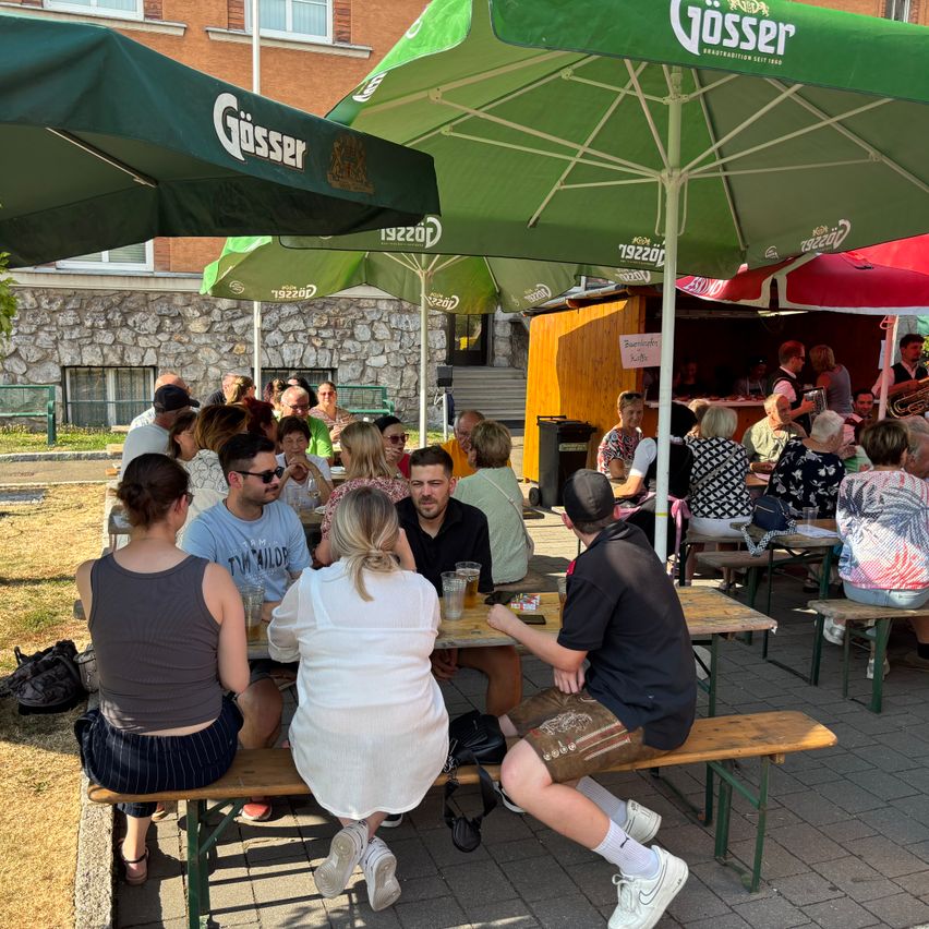 Eine Gruppe von Menschen sitzt unter grünen Gosser-Sonnenschirmen an Tischen. Die Tische haben Gläser mit Bier und Essen. Im Hintergrund steht ein Gebäude mit Fenstern und einem Imbissstand.