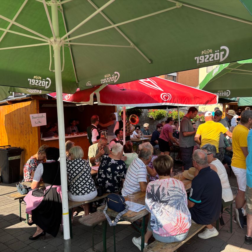 Menschen versammeln sich unter grünen und roten Schirmen auf einem Freimarkt. Einige sitzen an Tischen, andere stehen und unterhalten sich. Ein Verkäufer steht hinter einem Tresen, und andere gehen umher.