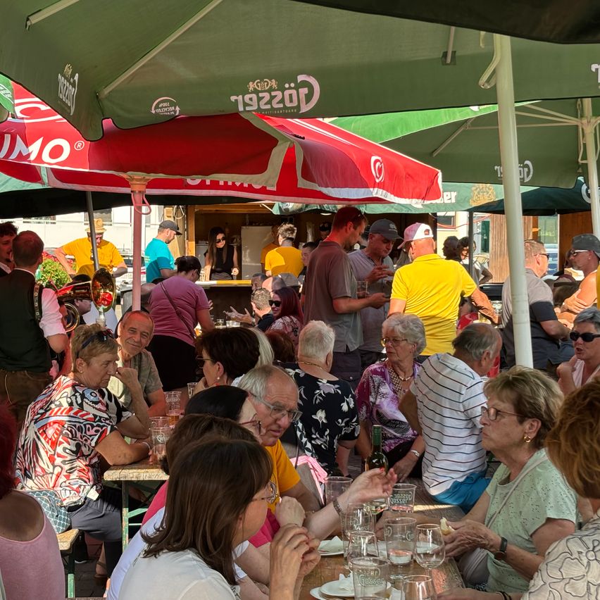 Ein Café im Freien mit vielen Menschen, die an Tischen unter roten und grünen Schirmen sitzen und stehen. Einige reden, andere essen und trinken. Auf den Tischen stehen Flaschen und Gläser.