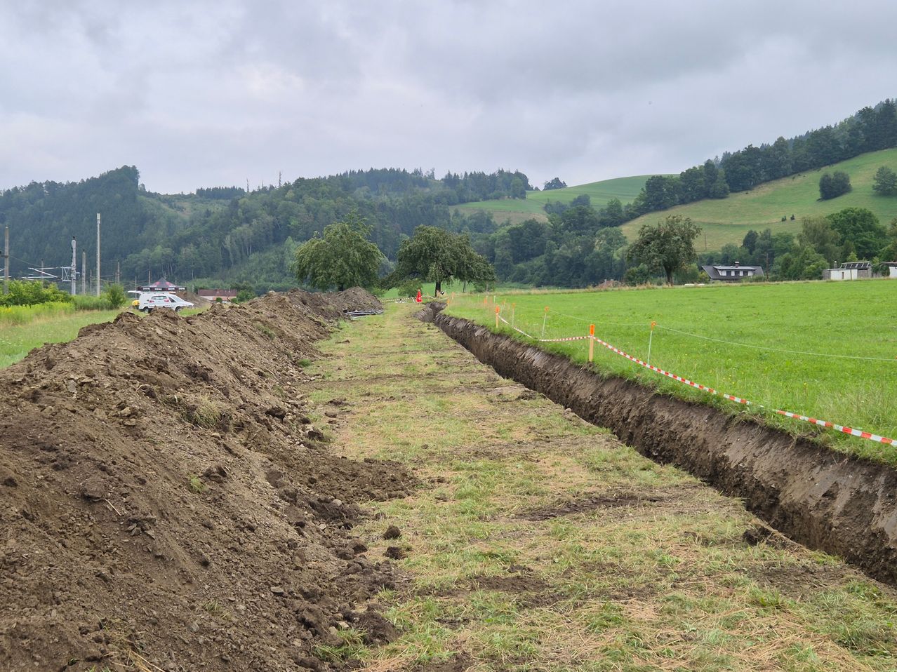 Eine Baustelle in einem ländlichen Gebiet, mit einer frisch gegrabenen Rinne, die durch ein saftiges Feld verläuft, wobei Bäume und ein Gebäude im Hintergrund unter einem bewölkten Himmel zu sehen sind.