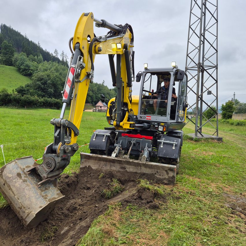 Ein gelber Bagger gräbt in die Erde, mit einem Mann am Steuer auf einem Rasen, einem Turm im Hintergrund und Bäumen und einem Haus in der Ferne.