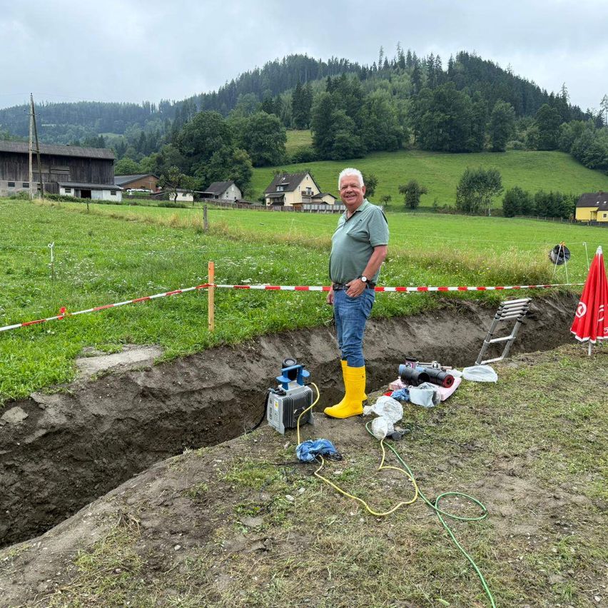 Ein Mann in gelben Stiefeln steht auf einem Feld mit einer großen Grube und Ausrüstung in der Nähe, mit Häusern und einem Bauernhaus im Hintergrund.