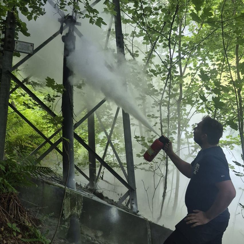 Ein Feuerwehrmann sprüht Wasser aus einem Feuerlöscher in einem Wald, umgeben von Bäumen und Nebel.