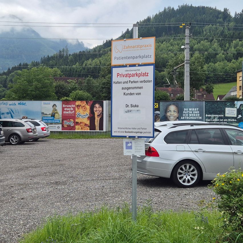 Ein Patientenparkplatz mit einem Verbotsschild, umgeben von Werbetafeln und Bergen. Mehrere Autos sind geparkt.