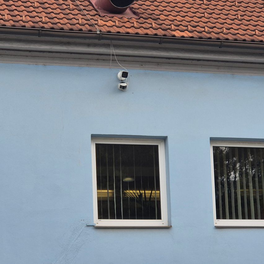 Ein blaues Gebäude mit einem Ziegeldach und zwei Fenstern, eines mit Gittern und das andere offen. Zwei Sicherheitskameras sind am Dach montiert.