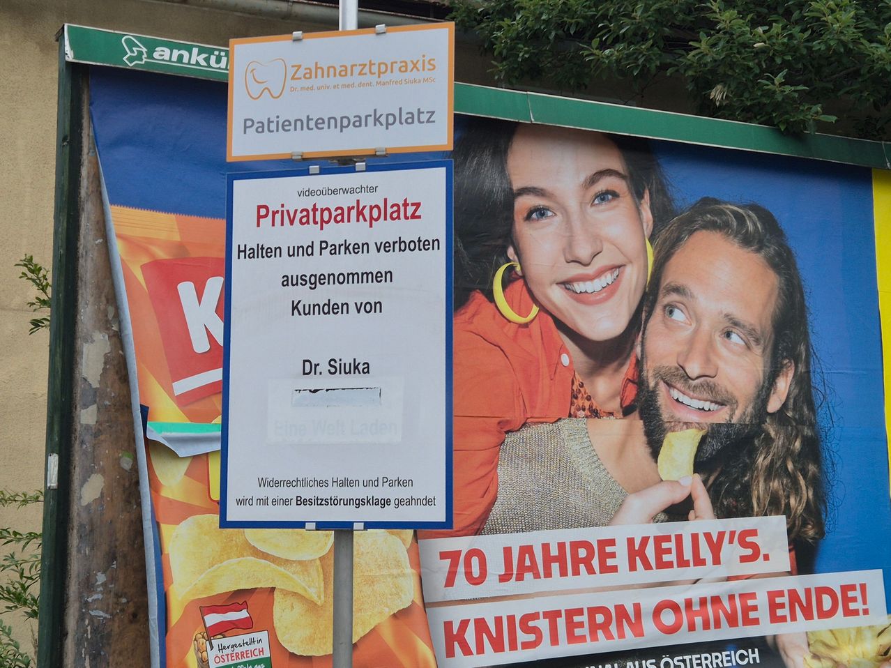 Ein Schild verbietet das Parken auf einem privaten Parkplatz für Patienten einer Zahnarztpraxis. Darunter befindet sich eine große Werbung für Kelly's Kartoffelchips mit einem lächelnden Paar, das Chips isst.
