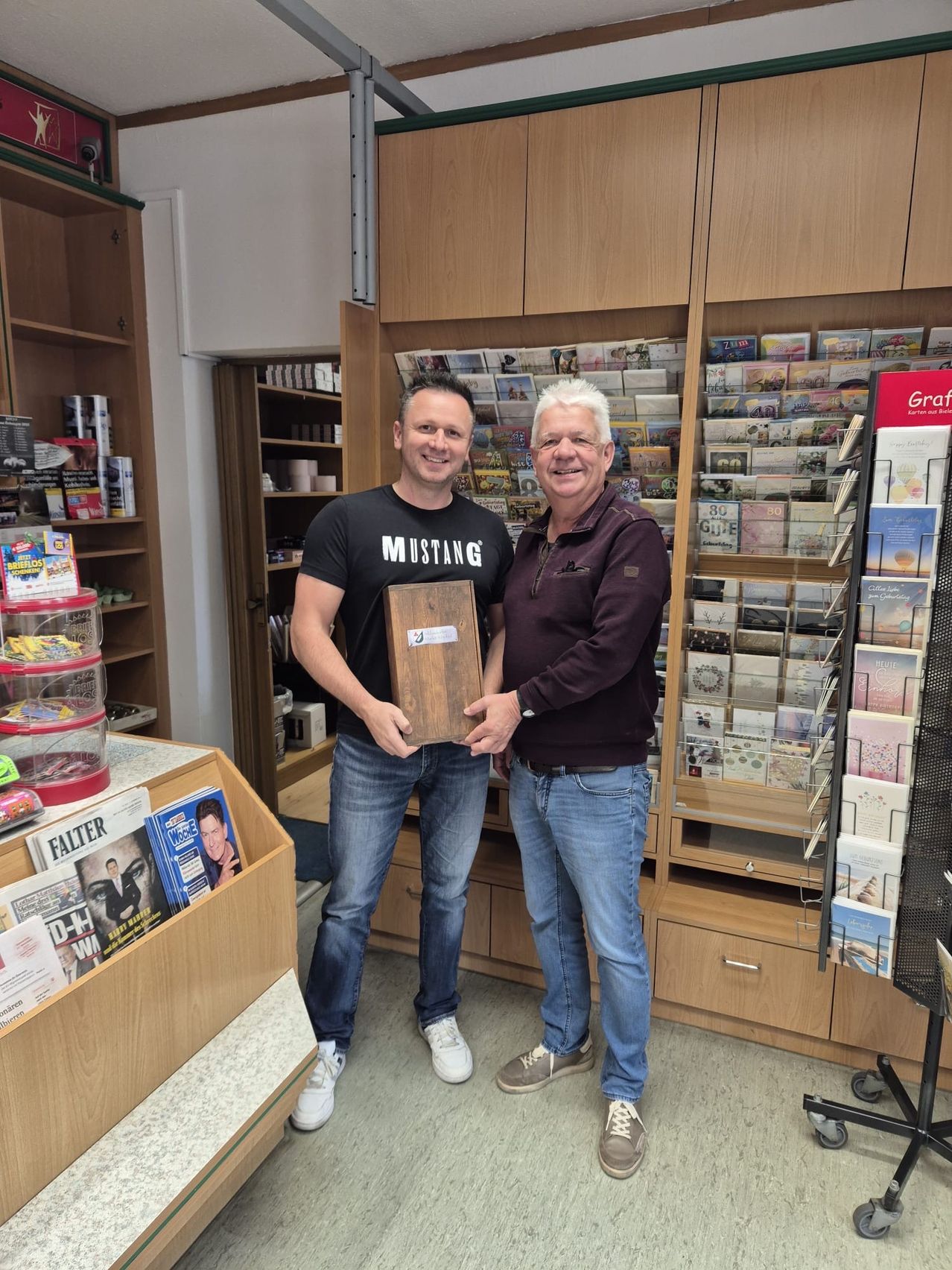 Zwei Männer stehen lächelnd in einem Geschäft. Der Mann auf der linken Seite hält eine Holztafel mit dem Wort Mustang. Hinter ihnen befinden sich mit Zeitschriften und Büchern gefüllte Holzregale.