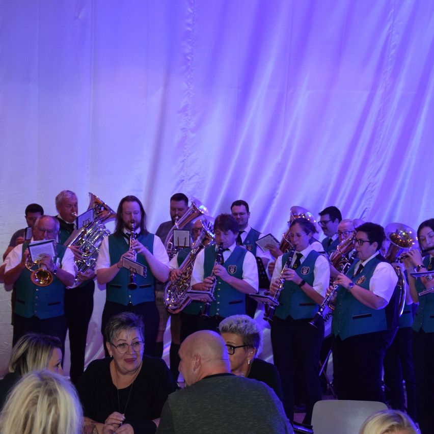 Ein Blasorchester spielt auf der Bühne, beleuchtet von lila Lichtern. Mehrere Musiker spielen verschiedene Instrumente, während einige Zuschauer davor sitzen.