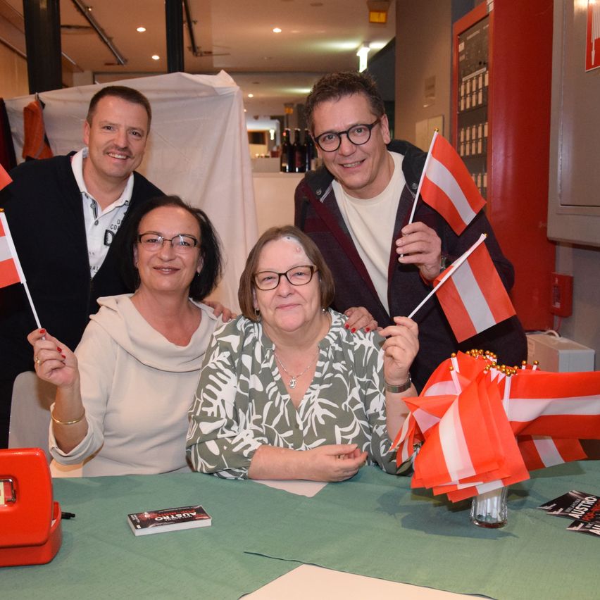 Vier Personen sitzen mit österreichischen Flaggen in der Hand an einem Tisch. Zwei Männer und zwei Frauen. Der Tisch hat ein grünes Tuch und verschiedene Gegenstände darauf. Dahinter steht eine weiße Struktur.