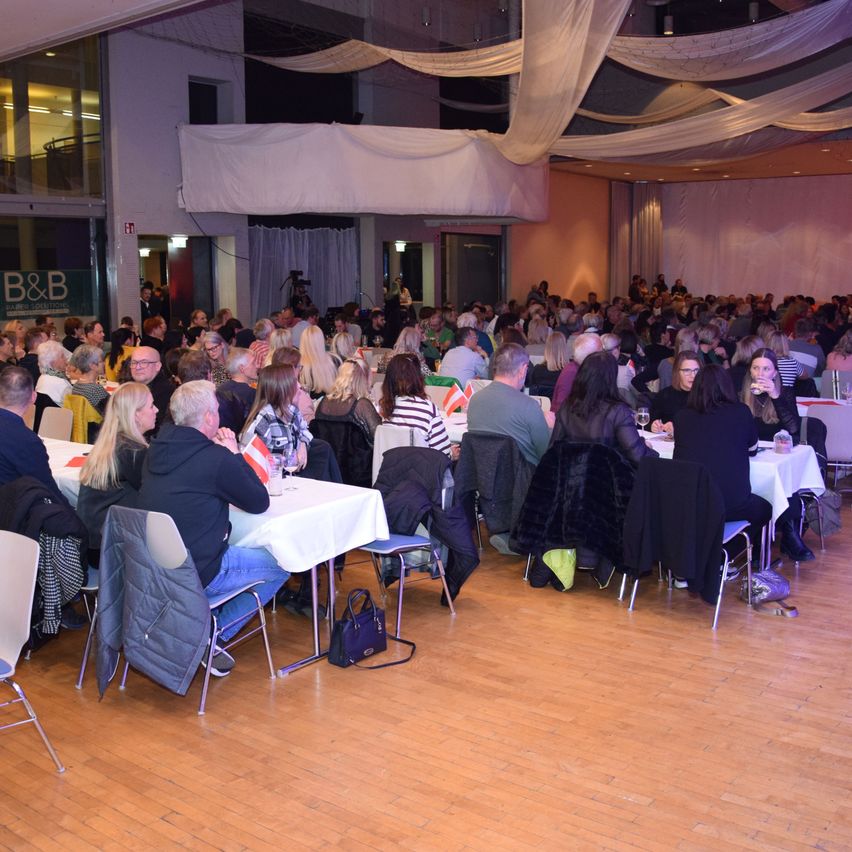 Eine große Gruppe von Menschen sitzt an Tischen in einem großen Raum, wahrscheinlich für ein Bankett oder eine Veranstaltung. Der Raum hat Holzböden und weiße Vorhänge. Einige Leute tragen Brillen und Hüte.