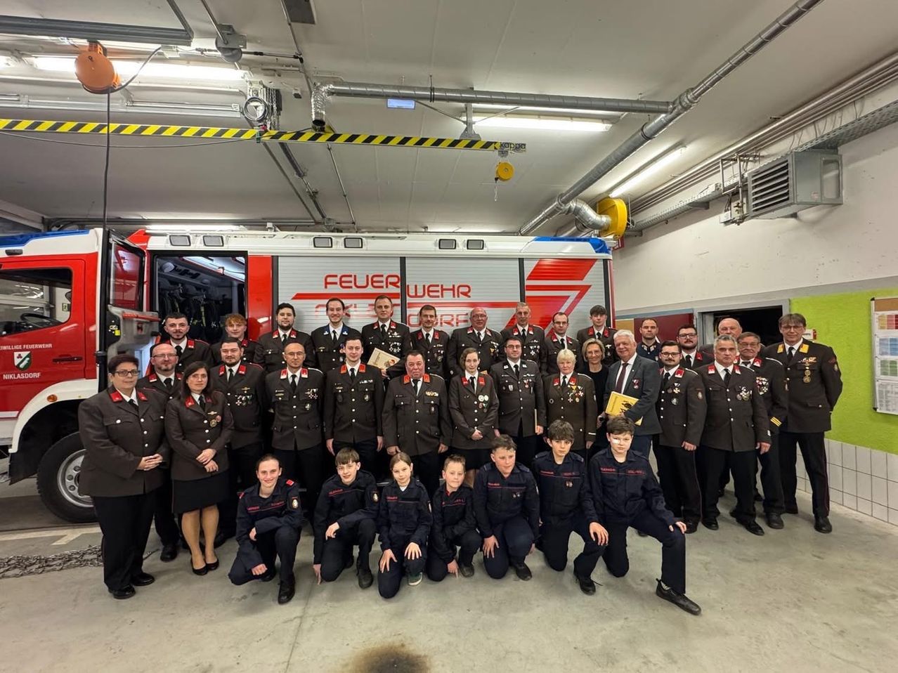 Eine Gruppe Feuerwehrleute in Uniform posiert für ein Foto in einer Feuerwache, mit einem Feuerwehrauto im Hintergrund.