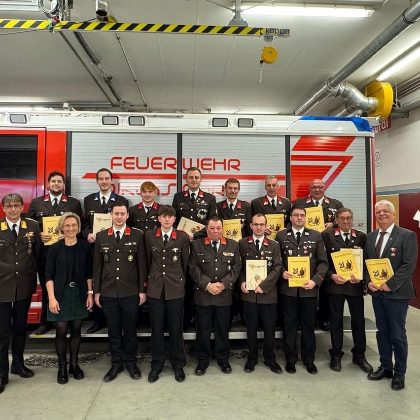 Eine Gruppe von Feuerwehrleuten, sowohl Männer als auch Frauen, posiert für ein Foto vor einem Feuerwehrauto und hält Urkunden in den Händen.