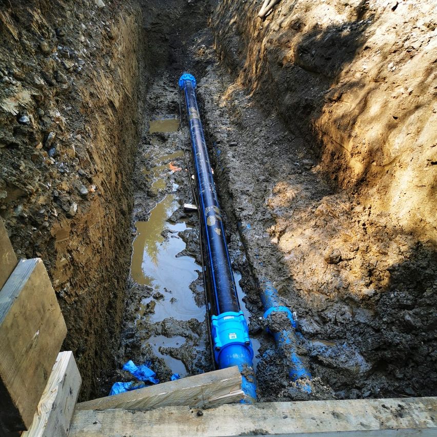 Eine große blaue Rohrleitung ist in einem mit schlammigem Wasser gefüllten Graben vergraben, mit Holzbrettern an den Seiten.