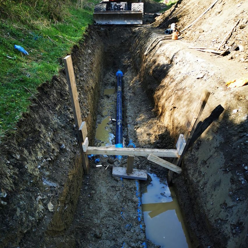 Eine Baustelle mit einer tiefen Grube und einer großen blauen Rohrleitung. Holzstützen halten die Ränder der Grube. Ein Bulldozer ist in der Nähe geparkt.