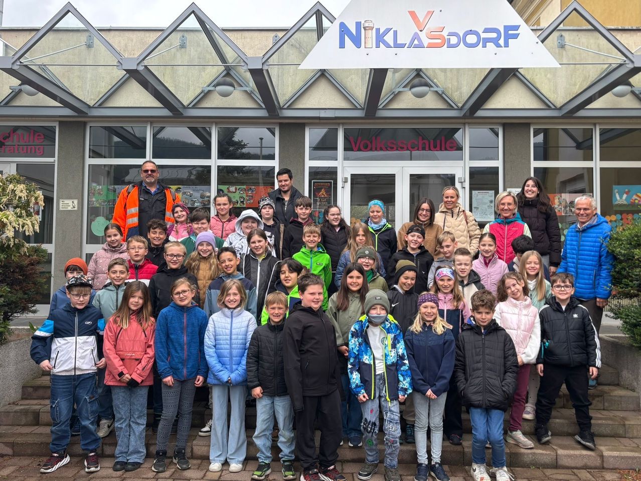 Eine Gruppe von Schülern und Erwachsenen posiert für ein Foto vor einem Gebäude mit dem Schild 'NIKLASDORF Volksschule'. Die Schüler tragen Winterkleidung, einige mit Masken, während zwei Erwachsene vorne eine Brille tragen.