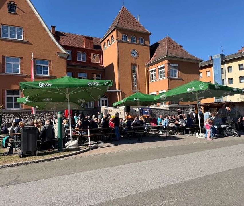 Ein Außenbereich zum Essen mit grünen Schirmen vor einem Gebäude. Viele Menschen sind versammelt, einige sitzen an Tischen und andere stehen. Ein Mülleimer und eine Fahne sind sichtbar.